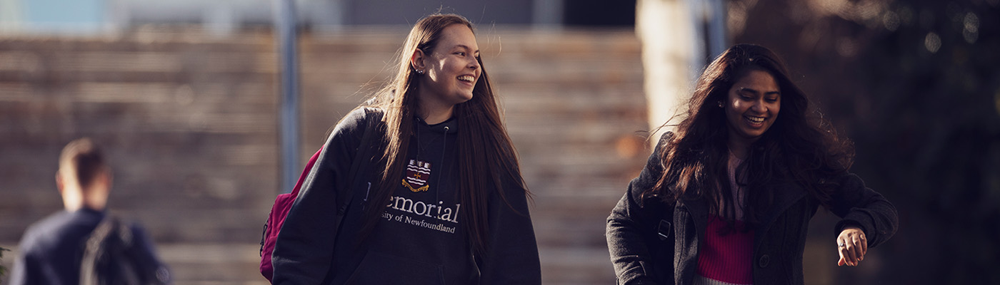 Two female university students, one Caucasion wth brown hair and one South Asian with brown hair, walking and smiling outdoors with stairs in the background.